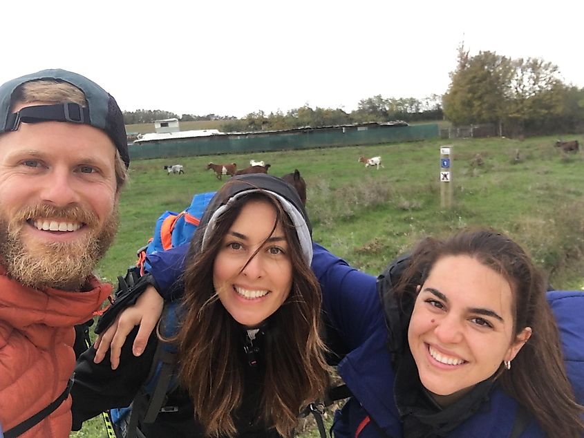 A man and two women happily pose in front of a grassy field full of goats. 